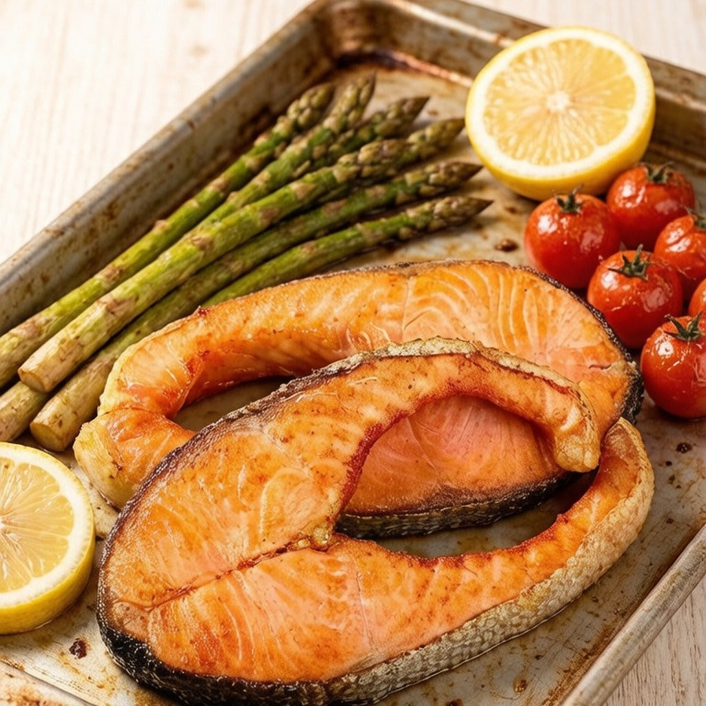 Two roasted salmon portions on a tray with asparagus, tomatoes and lemon